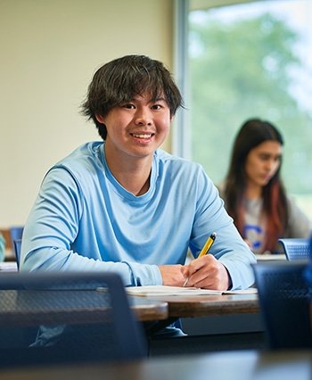 Male TCC student smiles in a classroom while taking notes.